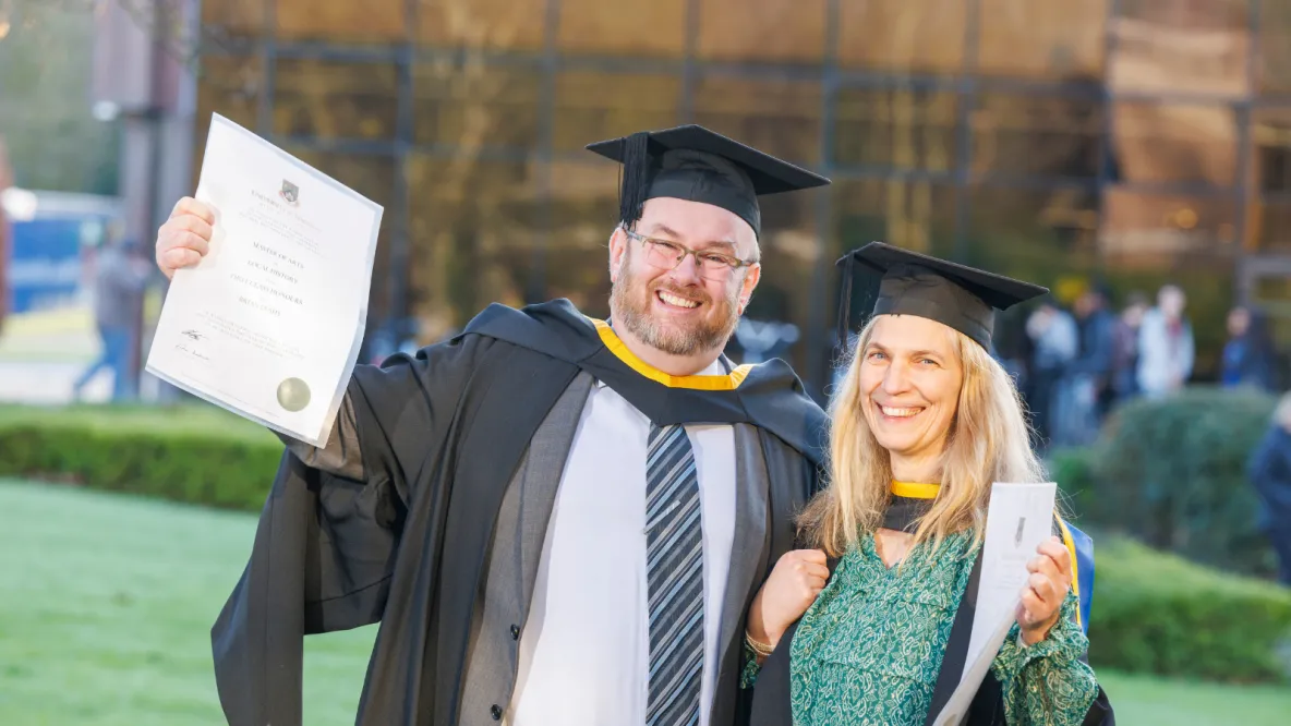 A photo of a man and woman in smart dress and graduation robes and cap, each holding up a degree certificate