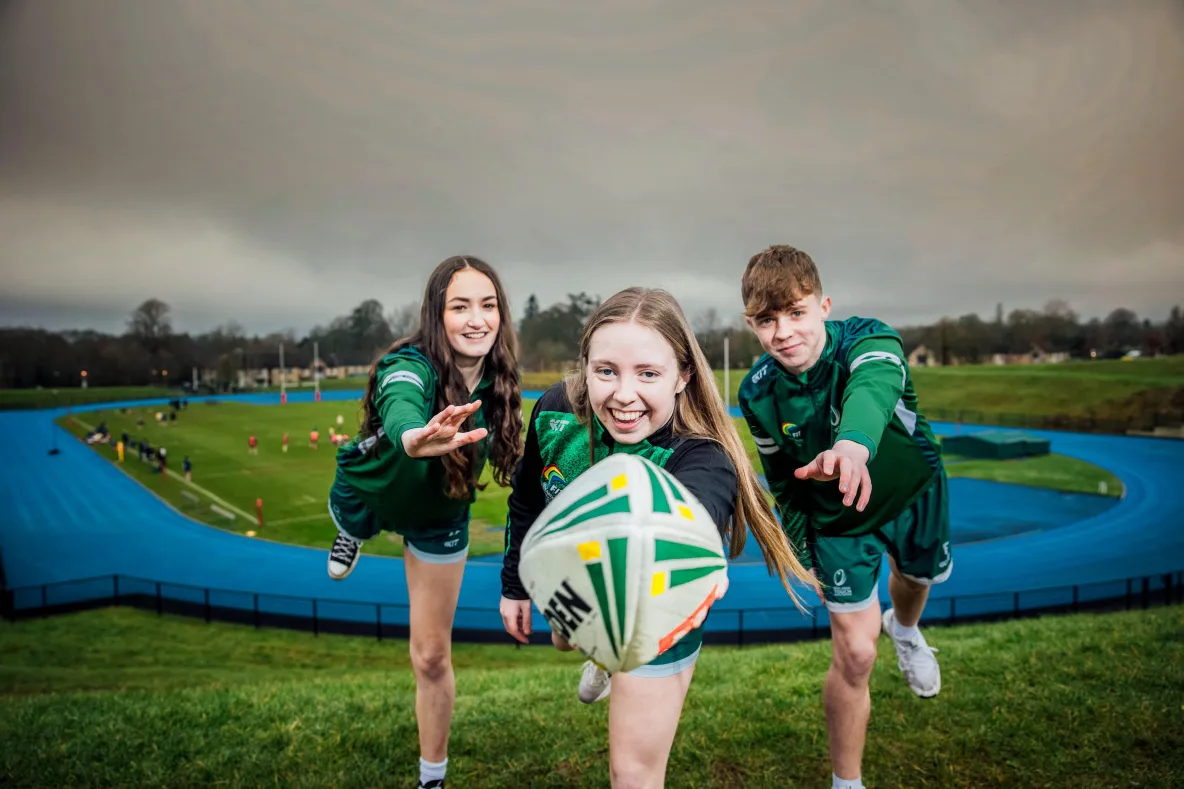 three youth touch rugby athletes, one male and two female, leaning towards camera with a rugby ball with a running track in the background