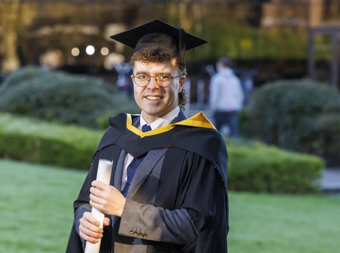 Man standing in outdoor setting wearing graduation robes and holding graduation scroll