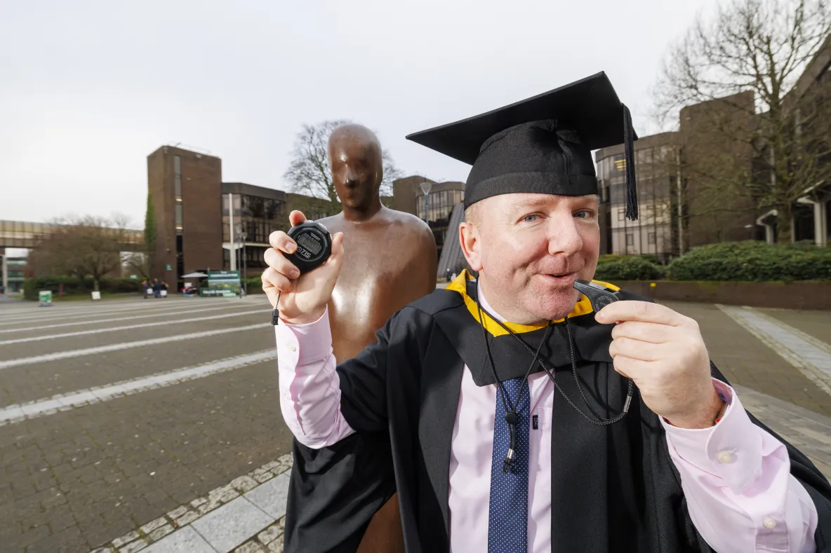 Man in gtraduation robes holding a coaches whistle and stopwatch standing in front a bronze statue with university building in the background