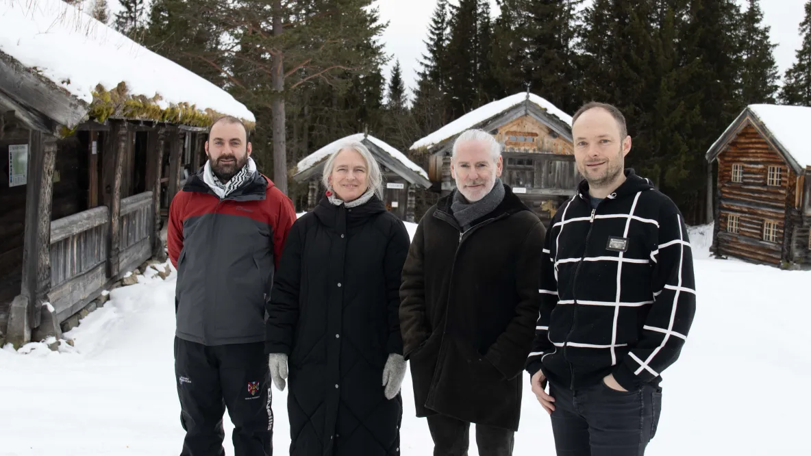 A photo of four people dressed in outdoor clothing, standing in snow in front of cabins and trees