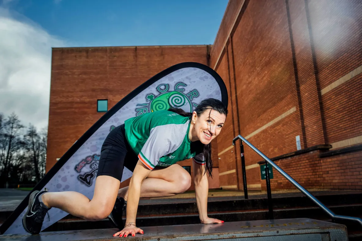 A female athlete with dark hair, wearing a green and white sports jersey, crouches on a metal platform, smiling at the camera. She appears to be mid-obstacle in an outdoor setting, with a brick building and a branded flag in the background. The flag displays a green spiral design with the words "OCR Ireland." The scene is set against a partly cloudy sky.