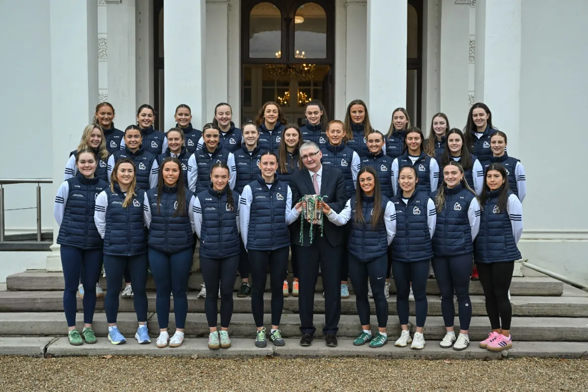 Acting President of the University of Limerick, Professor Shane Kilcommins, stands with the UL Ashbourne Cup-winning camogie team outside Plassey House. The team, dressed in navy UL jackets and white jerseys, proudly gathers around him as he holds the prestigious trophy. Smiling faces reflect their back-to-back championship success for UL camogie.