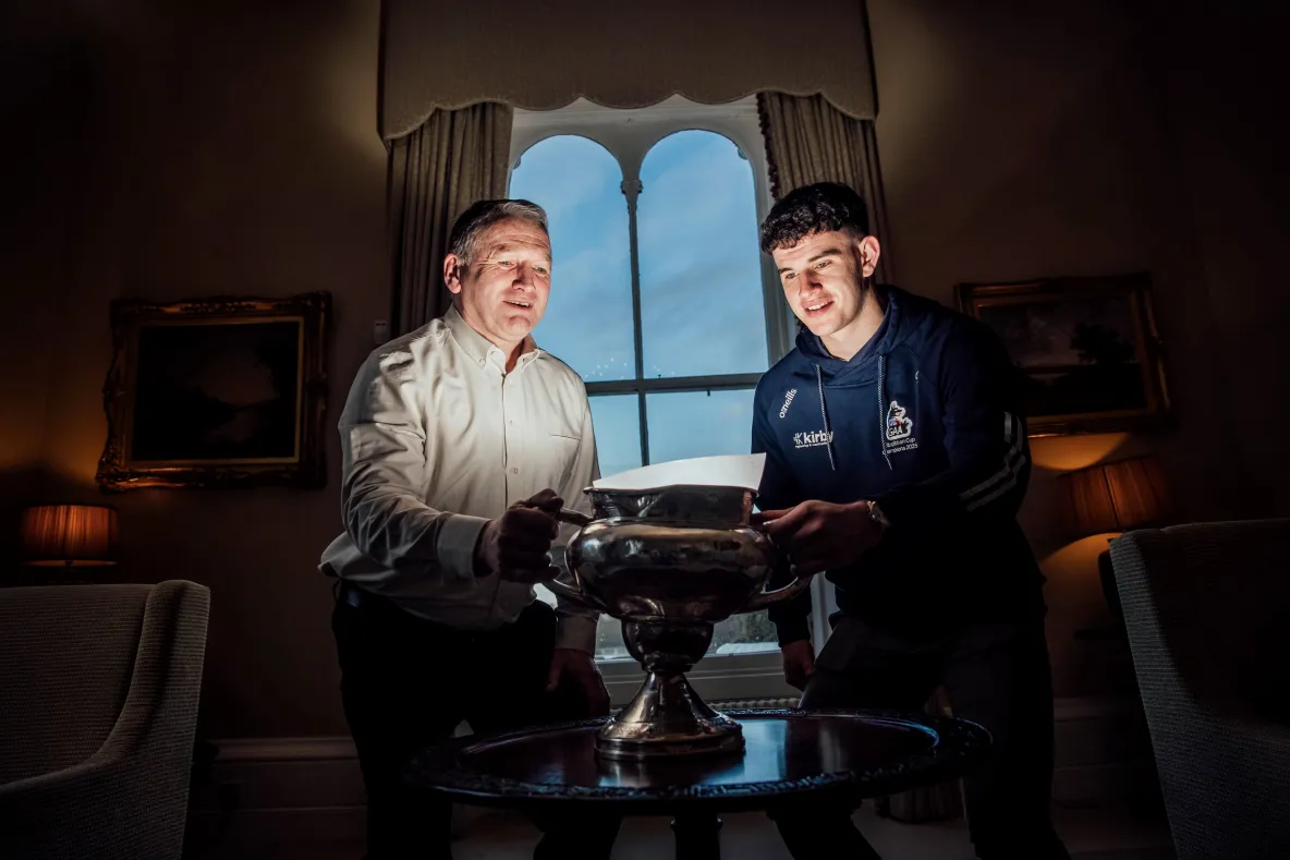 Two men, an older one to the left wearing a light shirt, and a younger man to the right wearing a navy hoody, in a stately setting, holding a champioship cup and looking into the bowl. There is a stately window behind them