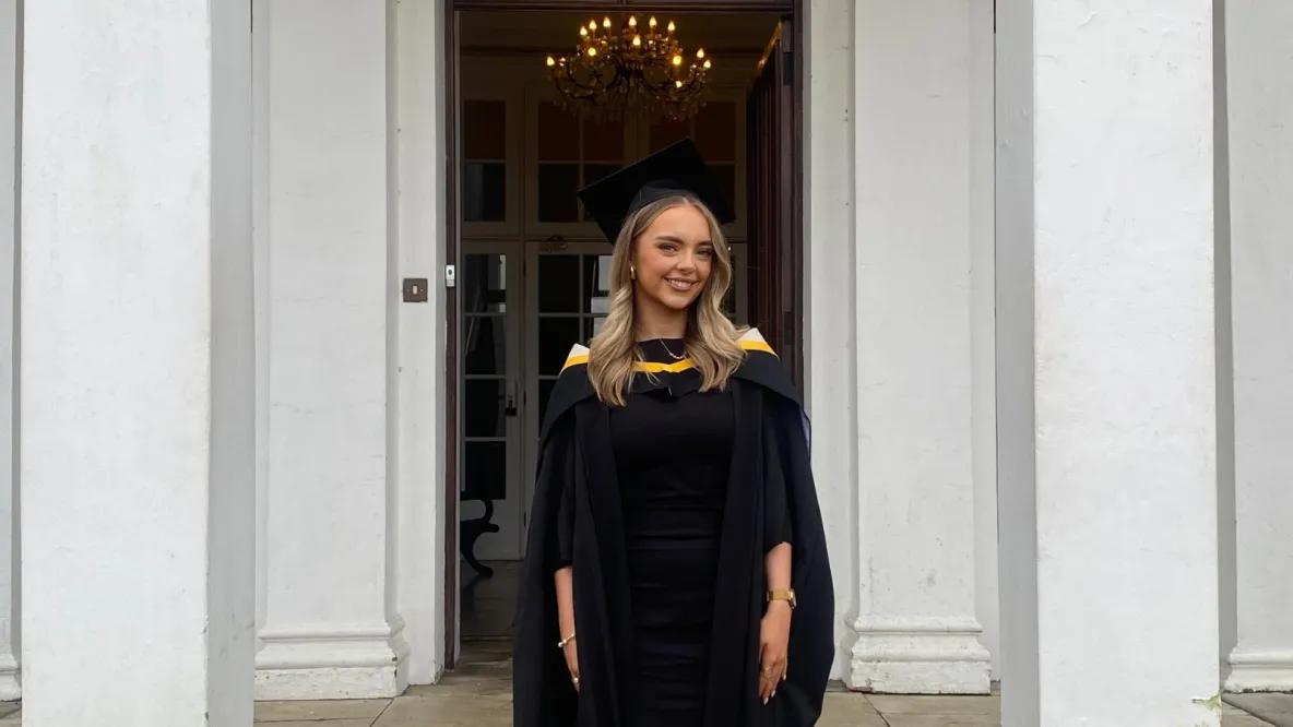 A photo of a woman in black graduate robes and cap, standing in front of a doorway