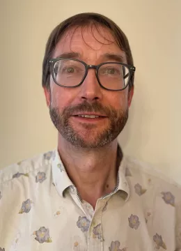 A man with dark hair and glasses standing and smiling