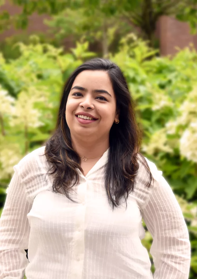 A young woman in a white top standing in front of flowers smiling