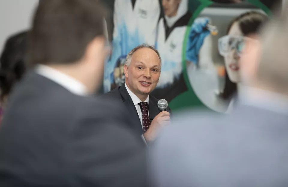 Man holding microphone speaking at conference Backs of two other men out of focus in foreground