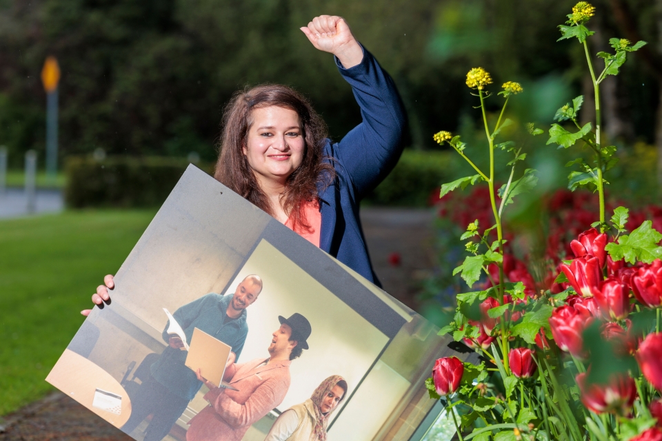 Woman celebrating with left arm aloft holding winning picture in right had, taken in garden with red flowers in foreground