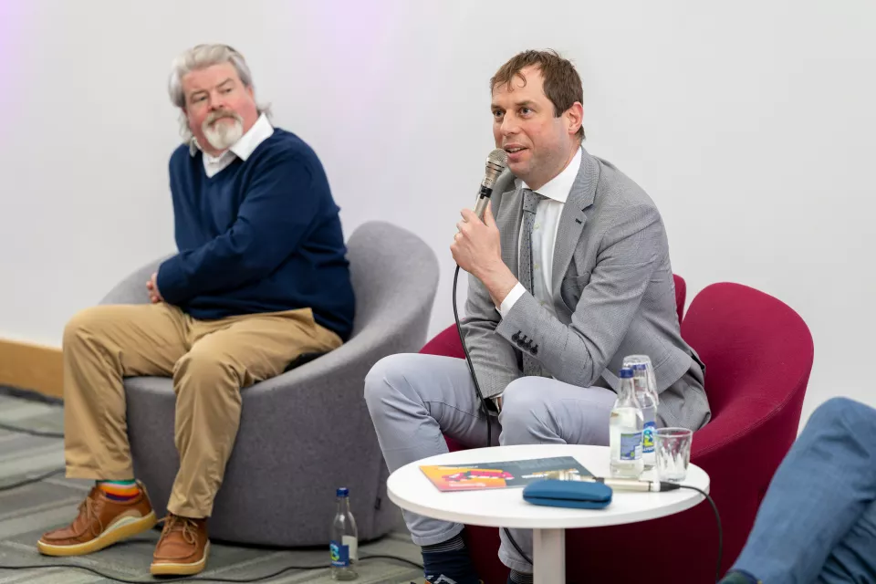 Two men sitting down, the man on right has a microphone and is speaking to an audience which is not in shot