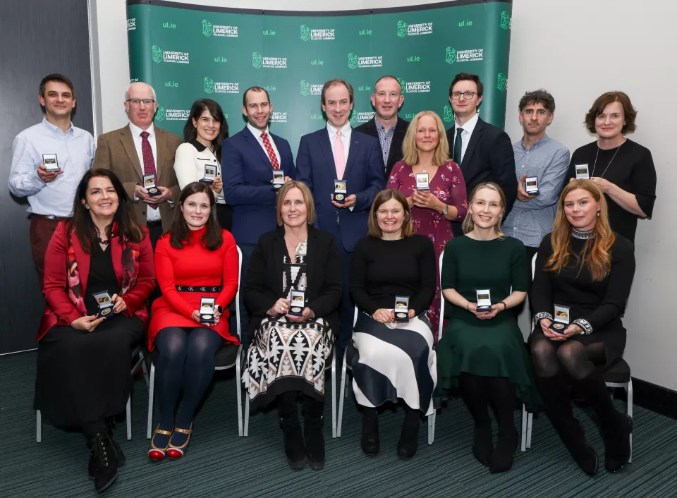 Group of nine women and seven men sitting and standing in front of UL backdrop with their medals for research excellence